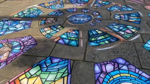 A colourful, spiralling artwork resembling a stained glass window has been created on paving slabs on the ground.