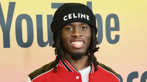 Getty Images Kai Cenat smiles towards the camera as he wears a black beanie hat and red letterman jacket.