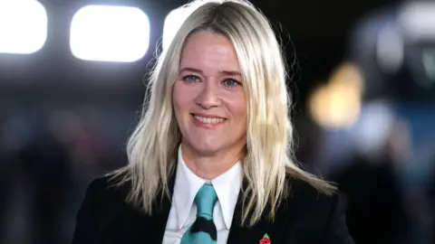 Getty Images Edith Bowman is wearing a black suit jacket, a white shirt and a blue tie. She is smiling, with blurred bright lights behind her.