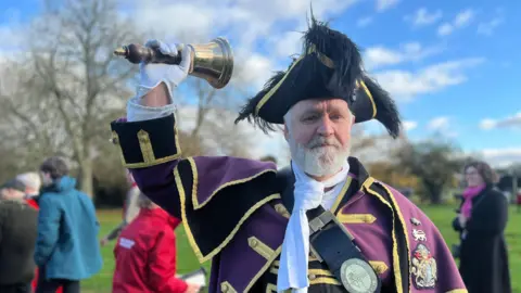 Royal Borough of Windsor and Maidenhead Town Crier Chris Brown with a white beard and wearing a purple and gold coat, a white scarf and a black feather hat. He is ringing a hand bell. It is a sunny day and there is a small group of people, some trees and a blue sky behind him.