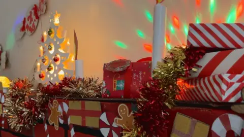 Alice Cullinane/BBC A brick fireplace is covered in gold and red tinsel and white candles. There are presents stacked on the right side of the fireplace with colourful lights in the background.