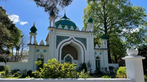 Alexa Rusakoff/BBC A white and green domed mosque stands in bright sunshine under a blue sky. There are ornate decorations and tiling on the mosque with trees and shrubs around the outside.