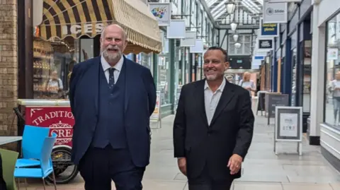 Bedford Borough Council Tom Wootton and John McReynolds walking down Bedford's Arcade shopping centre, wearing dark suits and smiling