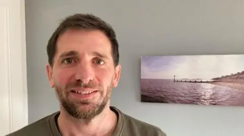 Head and shoulders of a man with short brown hair, and a short brown beard, standing in front of a plain backdrop, with half a slim painting of an ocean scene behind him. 