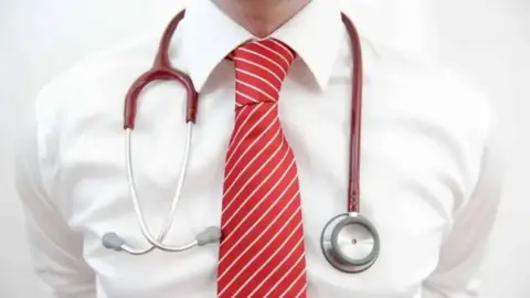 A close up of a man wearing a white shirt and red stripy tie, with a stethoscope around his neck.