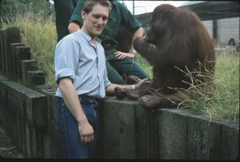 man with blue jeans and shirt posing with an orangutan who is sitting on a fence