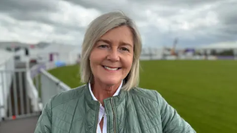 Rhonda has greyish, silvery hair that stops just above her shoulders. Behind her is a fenced off grassy area, which will probably be used to exhibit animals during Balmoral Show. 