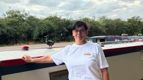 Shelton Pell A woman with short black hair and dark framed glasses stands with a narrowboat behind her, she is wearing a white t-shirt, a hedge is visible in the background.