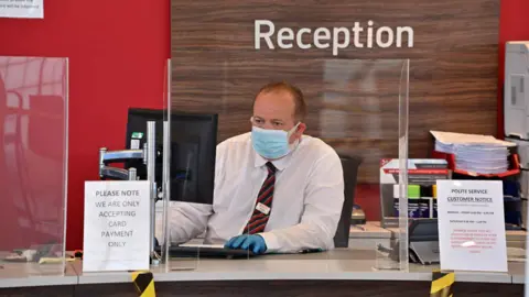 Getty Images An employee wears PPE (personal protective equipment) including a face mask and gloves as a precautionary measure against COVID-19, as they sit behind perspex screen in the reception of a recently re-opened Vauxhall car dealership in north London