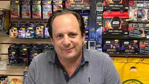 GUY CAMPBELL/BBC Manager of toy shop wearing striped blue top standing in front of a selection of games and toys 