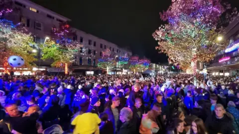 Johnny Rutherford/BBC A crowd of people watching the Plymouth city centre light switch-on. There is a blue glow over the crowd and trees are wrapped in colourful Christmas lights.