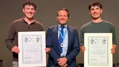Jordan Adams and his brother Cian are holding up certificates after being granted the Freedom of the Borough in Redditch. In between them is council leader Matt Dormer wearing a blue suit.