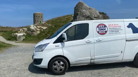 Lesley and Gary Ford A van from The Pet Bus parked up in a car park in the Channel Islands. The van is white and has a navy blue silhouette of a dog on the side alongside its logo and contact details. The van has the word Beau written on it near the front passenger door. A fortification is in the background.