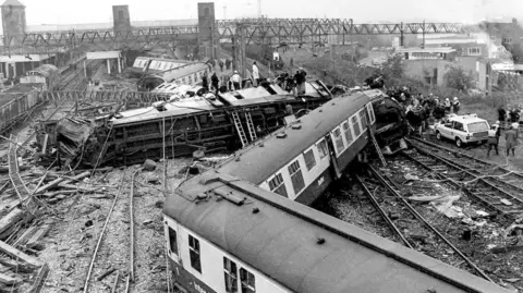 The wreckage after a train crash. Carriages zigzag across two railway tracks in this black and white image. The carriages are white with dark lower halves to their bodywork. One carriage is on its side in the distance while people mill around the wreckage.