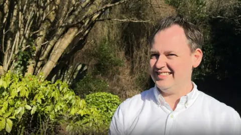 BBC Close up of Scott Bloomfield who is standing outside next to a road. Trees and bushes are behind him. Mr Bloomfield is wearing a white shirt and is smiling at the camera. 