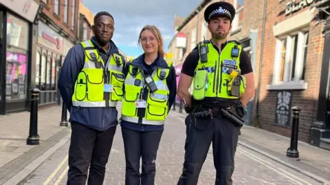 Standing in a cobbled street lined with shops, two men and a women stand side by side, looking straight on. They're all dressed in official florescent police officer vests, with walkie-talkies, official badges and lapel cameras.