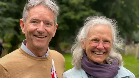 Big smiles from Charles Flinders Lucas who is wearing a sandy coloured jumper over a shirt and Aveley Lucas, wearing glasses, a purple scarf and denim jacket 