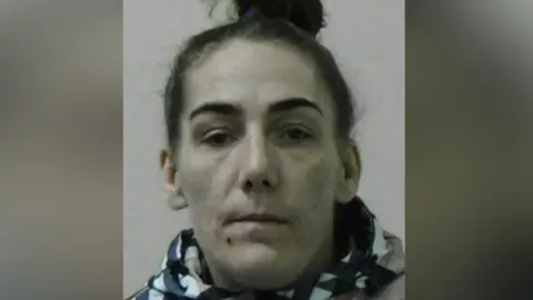 Mugshot of Victoria Pedersen. She is pale with a large red spot on her chin, and dark hair tied in a bun on top of her head