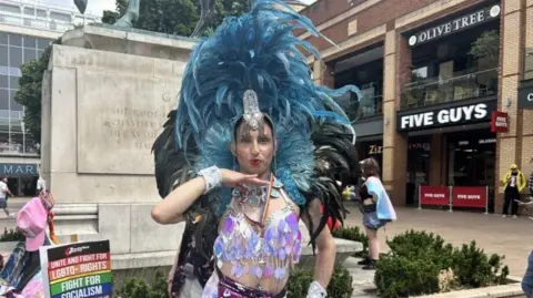 A drag performer in feathered purple bodice, silver necklace and ornate blue and black feathered headdress poses with their hand under their chin at the base of a stone plinth