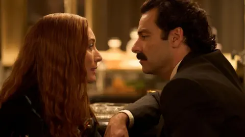 Robert Viglasky/Disney+ A shot from the series Rivals. It depicts the characters of Maud and Declan O'Hara looking at each other. Maud has long red hair and Declan has thick dark brown hair and moustache. 
