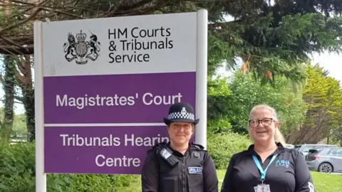 Sgt Alice McKinnon (left) and Kay Smith, anti-social behaviour officer for Camborne at Cornwall Council, stand next to a purple and white sign for Truro Magistrates' Court.
