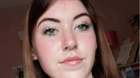 A teenage girl with should length, brown hair. She has a pale complexion and is wearing a nose ring.