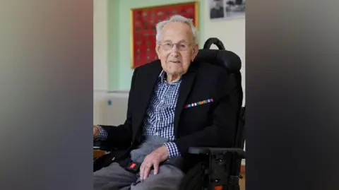 Ron Parker is in a wheelchair wearing grey trousers and a blue and white gingham check shirt. He has a black blazer bearing a bar of colours by the left lapel, which would typically signify the wearer has won war medals