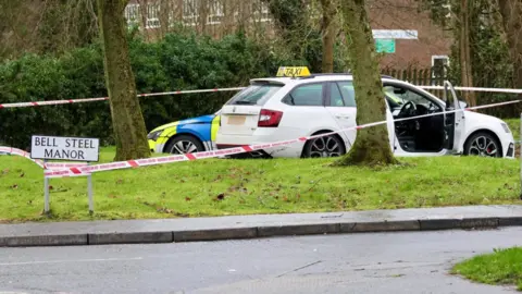 Pacemaker A white estate car with a taxi sign sits in a car park. There is a police cordon tied to a street sign for "Bell Steel Manor". A marked police car is parked behind the taxi. There are a variety of trees and patches of grass at the perimeter.
