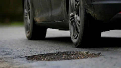 A large pot hole in a tarmac road with a black car driving past.