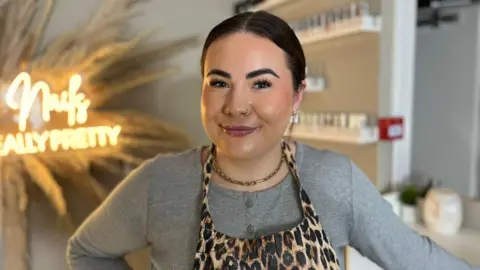 A woman with dark hair pulled back and a nose ring wears a leopard skin patterned apron over a grey top
