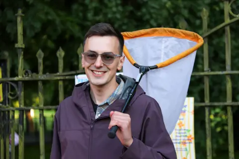 BBC / JOSH PARRY Entomologist Conor Butler holding an insect net over one shoulder, standing outdoors in front of a black metal fence and greenery