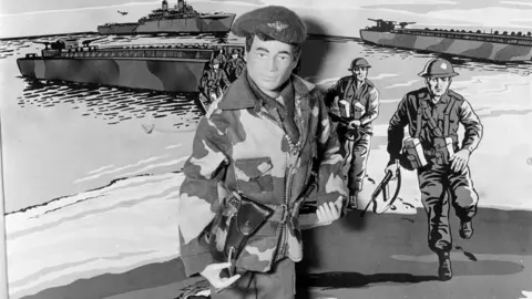 Mirrorpix/Getty Images A black and white image of an Action Man figure wearing military camoflauge and beret. He is stood in front of a pop art background depicting soldiers arriving on a beach by boat.