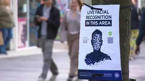 A sign posted to a tree saying 'facial recognition in this area' as people walk past in a street behind it