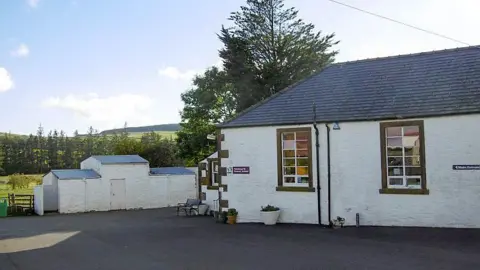 Richard Webb A small whitewashed rural school building, single-storey with white outbuildings and tarmac in front with trees and hills in the distance