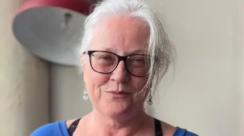Majella Berrnad is wearing black-rimmed glasses and a blue top.
She has long hair and is wearing earrings.
She is standing in front of a large red lamp.