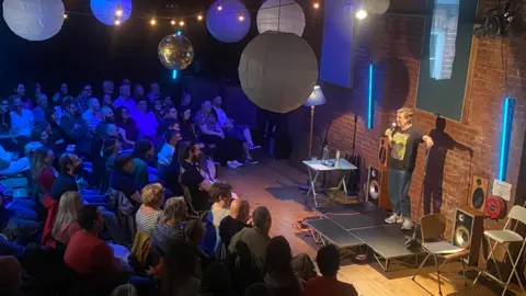 Josie Long, dressed in jeans and a black T-shirt, holding a microphone while stood on stage in front of a crowd.