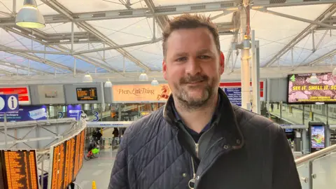 Photograph of Simon Bennett, the station manager at Manchester Piccadilly Station. The image shows the departure board and the platforms in the background.