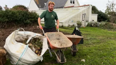 BBC Jack Etheridge, wearing a green T-shirt, pushing a wheelbarrow beside a bag of green waste