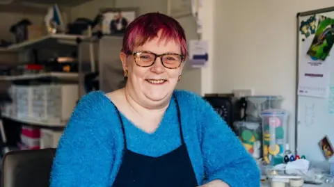 Gillian Colley Gillian Colley smiles at the camera as she is photographed in her workshop. She has a blue jumper on and black pinafore. She has pink hair and glasses. 