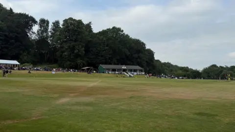 Nisha Joshi/BBC The cricket ground which is large expanse of cut grass with a green, small single-storey building on top of a small hill. A crowd of people can be seen sitting on either side of the building. There are trees surrounding the pitch and the sky looks grey.