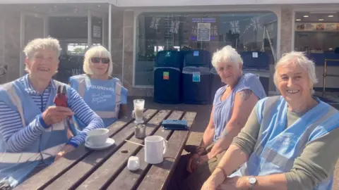 Claire Hamilton/BBC Four older ladies sit at a wooden outdoor table. They are each wearing blue vests that say 'volunteer'. Lady on the left is holding up a pair of secateurs.