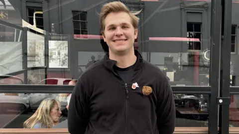 Edward stands in front of a window smiling. He wears a black jumper and has blonde hair. 
