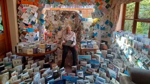 Kineton Manor Nursing Home An old man in a white shirt and black trousers holds a card in a room that is filled with birthday cards - on the floor and covering every inch of the walls