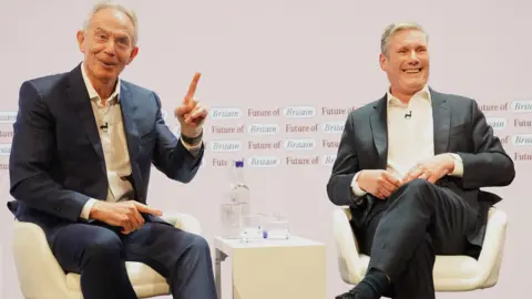 PA Tony Blair is sat to the left of Keir Starmer. Blair wears a navy suit and is holding his left hand in the air, pointing his finger. Keir Starmer is sat next to him. Both are laughing and appear to be in good spirits.