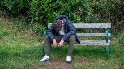 Getty Images A man sits on a bench in a grassy area with a hedge behind. He is hanging his head and wearing a blue coach with green tracksuit bottoms and trainers.