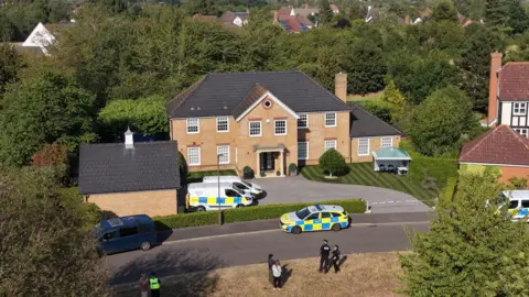 Ant Saddington/BBC Drone view of the house in Carnoustie Drive on a bright sunny day showing police activity and vehicles. The house is a large detached property with a pillared front entrance and sweeping drive with neat lawns and sheds
