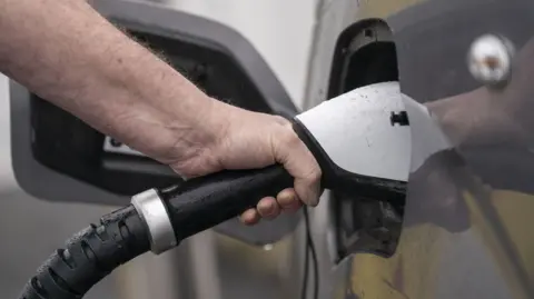 A close-up of a hand holding an electric car charger in the car socket. It looks like a dark-coloured car.