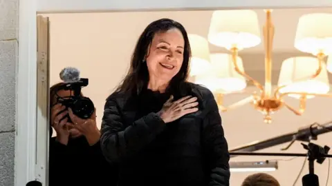 Nobel Peace Prize laureate Maria Corina Machado stands on a balcony with photographers and a hanging light fixture in a hotel room behind her. It is nighttime and she has her hand over her heart. She is smiling and wearing dark clothing.