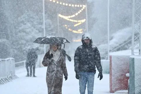 Reuters A woman, wearing a brown furry coat and cream scarf, while carrying an umbrella and a man wearing a black puffer jacket and jeans are covered in snow. Snow lying on the ground and falling from the sky is visible in the backround