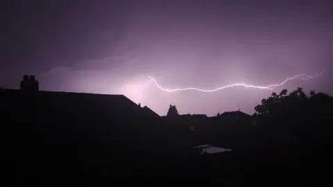 Josh/Weather Watchers A single bolt of lightning just above the silhouette of houses in a purple sky